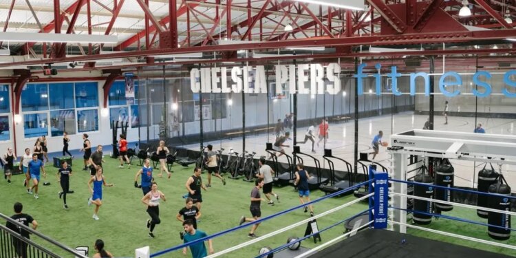 An indoor fitness facility with people exercising on a green turf, a boxing ring, and a basketball court, with "Chelsea Piers Fitness" written overhead.