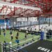 An indoor fitness facility with people exercising on a green turf, a boxing ring, and a basketball court, with "Chelsea Piers Fitness" written overhead.