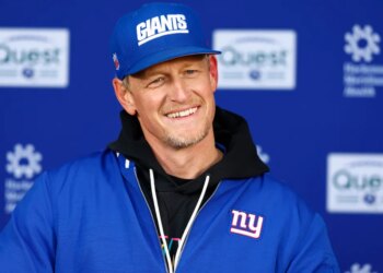 New York Giants defensive coordinator Charlie Bullen smiles while wearing a blue Giants hat and jacket during football practice.