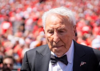 ESPN analyst Lee Corso wearing a black suit and bow tie, with an American flag lapel pin, leaving the field after his final pick.