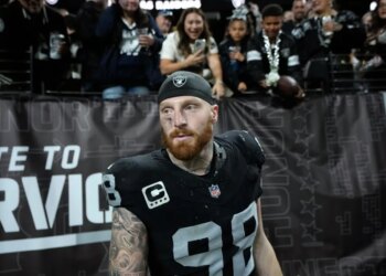A Dallas Cowboys football player in a black jersey with number 98, a black skull cap, and a red beard looks ahead, as fans watch from behind a fence.