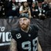 A Dallas Cowboys football player in a black jersey with number 98, a black skull cap, and a red beard looks ahead, as fans watch from behind a fence.