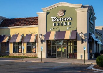 Panera Bread storefront with a beige and green facade, dark and light yellow awnings, and an empty parking lot on a sunny day.