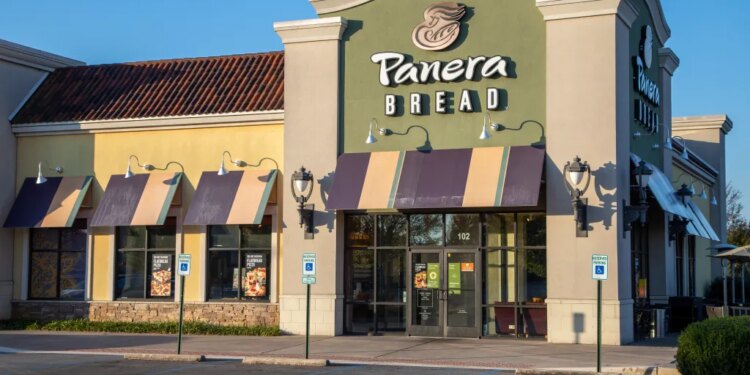 Panera Bread storefront with a beige and green facade, dark and light yellow awnings, and an empty parking lot on a sunny day.