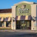 Panera Bread storefront with a beige and green facade, dark and light yellow awnings, and an empty parking lot on a sunny day.