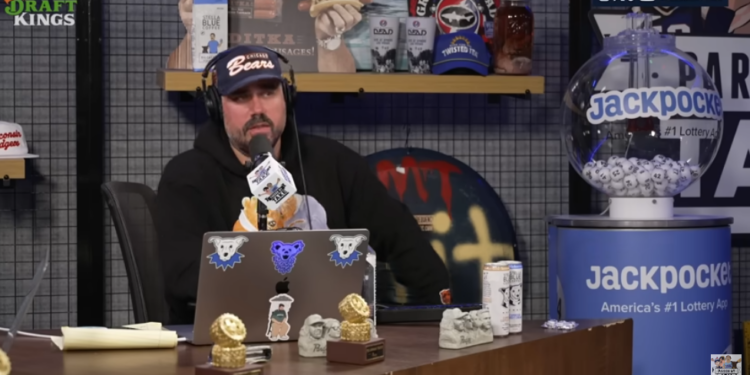 Katz sitting at a desk with a microphone and laptop, next to a lottery machine.