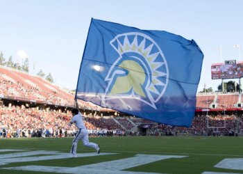A San Jose State Spartans flag bearer runs with a large blue flag with a yellow and white Spartan logo across a football field.