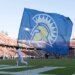 A San Jose State Spartans flag bearer runs with a large blue flag with a yellow and white Spartan logo across a football field.