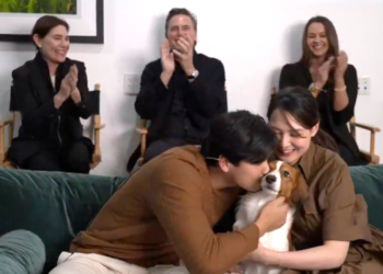 A couple sitting on a couch, with a man kissing a brown and white dog, and a woman smiling next to them, while three people in the background clap.