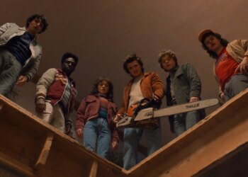 Six teenagers standing together on a ledge within a building. The teens are all looking down at something in confusion.