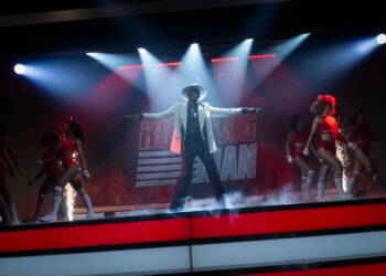 A man in a white jacket, white hat, and black pants dancing on a stage with a group of women dressed in bikinis, briefs, and knee high socks.