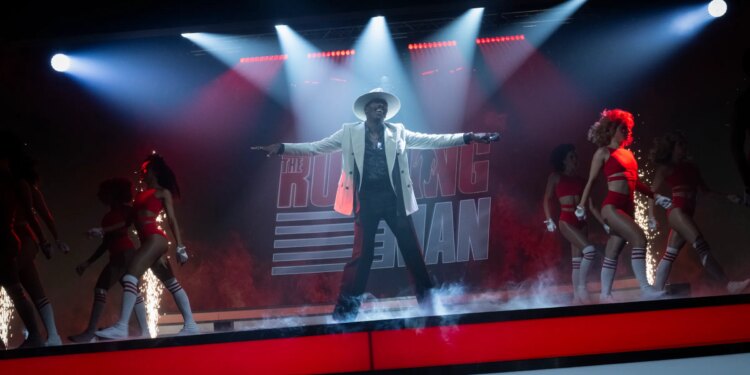 A man in a white jacket, white hat, and black pants dancing on a stage with a group of women dressed in bikinis, briefs, and knee high socks.