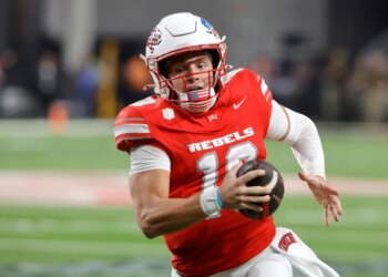 UNLV football player with number 10, wearing a red jersey, white helmet, and American flag chinstrap, running with the ball.