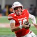 UNLV football player with number 10, wearing a red jersey, white helmet, and American flag chinstrap, running with the ball.