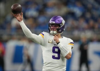 A Minnesota Vikings quarterback wearing a purple helmet and white jersey with the number 9, holding a football up in the air.