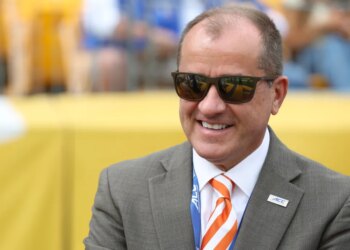 Man in sunglasses, suit, and orange striped tie, smiling at a football game.