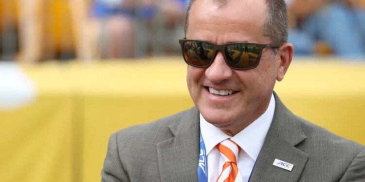 Man in sunglasses, suit, and orange striped tie, smiling at a football game.