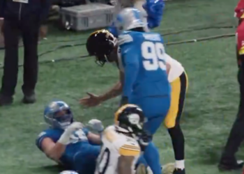 A football player in a blue jersey with number 99 is standing over another player on the ground in a blue jersey, while a third player in a black and yellow jersey is also on the ground.