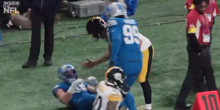 A football player in a blue jersey with number 99 is standing over another player on the ground in a blue jersey, while a third player in a black and yellow jersey is also on the ground.