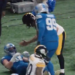 A football player in a blue jersey with number 99 is standing over another player on the ground in a blue jersey, while a third player in a black and yellow jersey is also on the ground.
