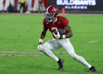 A football player in a red jersey and white pants running with the ball on a green field.
