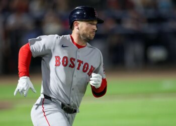 A Boston Red Sox player wearing a helmet and a grey uniform with "BOSTON" written in red on the front, running on a green baseball field.