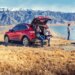 A red 2020 Ford Escape with its rear hatch open, with two women sitting on the bumper drinking from mugs, and a man fishing from a lake with mountains in the background.