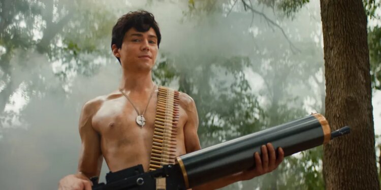Miles Heizer as Cameron Cope, shirtless with an ammunition belt over his shoulder and a large weapon, in a misty forest.