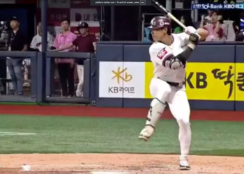 A baseball player in a white uniform with a maroon helmet and a bat in hand, standing at home plate, with "KB" and "KB LIFE" visible on the surrounding signage.