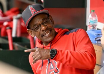 Ron Washington in a red hoodie and Los Angeles Angels cap, smiling and pointing in the dugout.