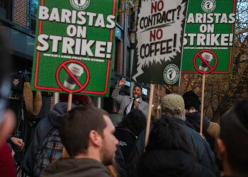 A man speaks into a microphone at a Starbucks union strike, surrounded by picketers holding signs that read "BARISTAS ON STRIKE!" and "NO CONTRACT? NO COFFEE!"