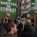 A man speaks into a microphone at a Starbucks union strike, surrounded by picketers holding signs that read "BARISTAS ON STRIKE!" and "NO CONTRACT? NO COFFEE!"