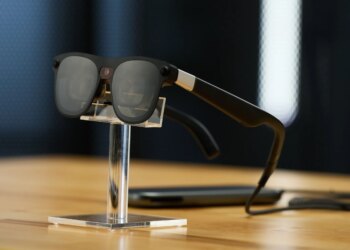 Close up of Project Aura glasses on a stand. You can see that they are wired and connect to a puck. They look like sunglasses, but in the nose bridge there is a camera.