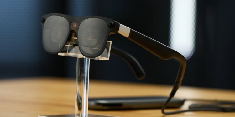 Close up of Project Aura glasses on a stand. You can see that they are wired and connect to a puck. They look like sunglasses, but in the nose bridge there is a camera.