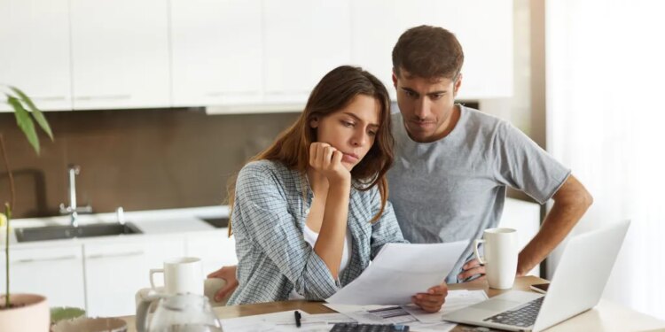 A young couple looking worried while reading financial documents in their kitchen.