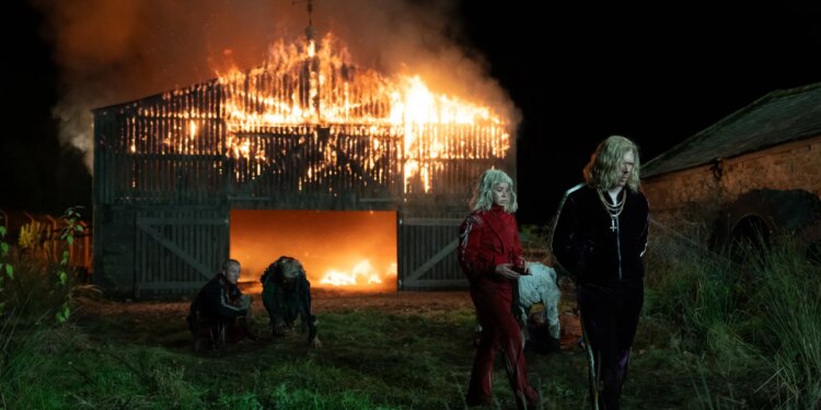 A man and a woman wearing platinum blonde wigs and track suits. The pair are walking away from a burning barn. Behind the two are two more people in wigs and track suits.