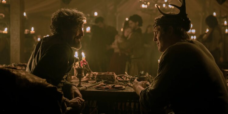Two men sitting together at a table in room where a party is being held. The man on the right is wearing a crown adored with antlers.