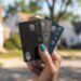 A hand holding three Bilt credit cards in black, silver, and blue, against a blurred suburban background.