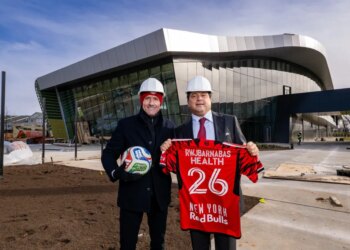 Two men in hard hats pose outside the Red Bull New York Training Facility, one holding a soccer ball and the other a New York Red Bulls jersey.