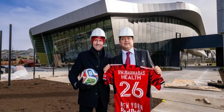 Two men in hard hats pose outside the Red Bull New York Training Facility, one holding a soccer ball and the other a New York Red Bulls jersey.