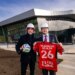 Two men in hard hats pose outside the Red Bull New York Training Facility, one holding a soccer ball and the other a New York Red Bulls jersey.
