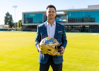UCLA Football Head Coach Bob Chesney holding a gold helmet with "UCLA" written on it in blue.