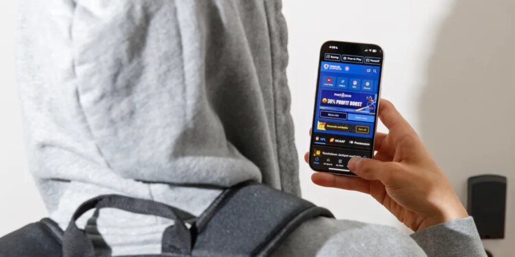 A person wearing a grey hoodie and backpack uses the FanDuel Sportsbook app on their phone.