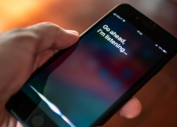 A hand holding an iPhone with the Siri interface showing the text "Go ahead, I'm listening..."