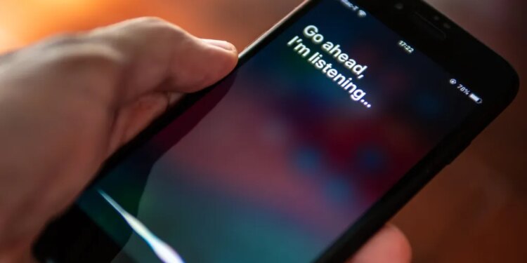 A hand holding an iPhone with the Siri interface showing the text "Go ahead, I'm listening..."
