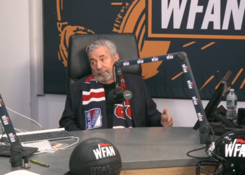 A man wearing a New York Rangers scarf sits at a desk with a WFAN microphone, a basketball, and a football helmet, all branded with WFAN.