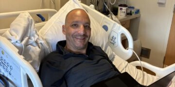 A man smiling from a hospital bed while using a laptop.