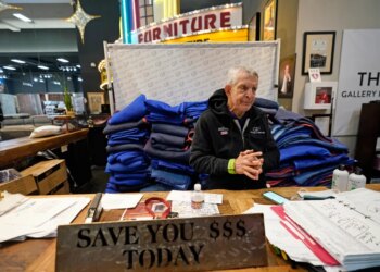 Jim McIngvale, owner of Gallery Furniture, talking at a counter in his store, with a sign that reads "SAVE YOU $$$ TODAY."