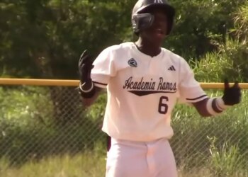 Baseball player Wandy Asigen in a white uniform and helmet, with "Academia Ramos 6" on his back, gesturing excitedly.