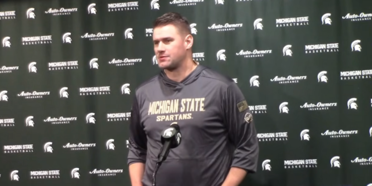 A man in a Michigan State Spartans hoodie stands in front of a green backdrop with repeated logos for Michigan State Basketball and Auto-Owners Insurance.